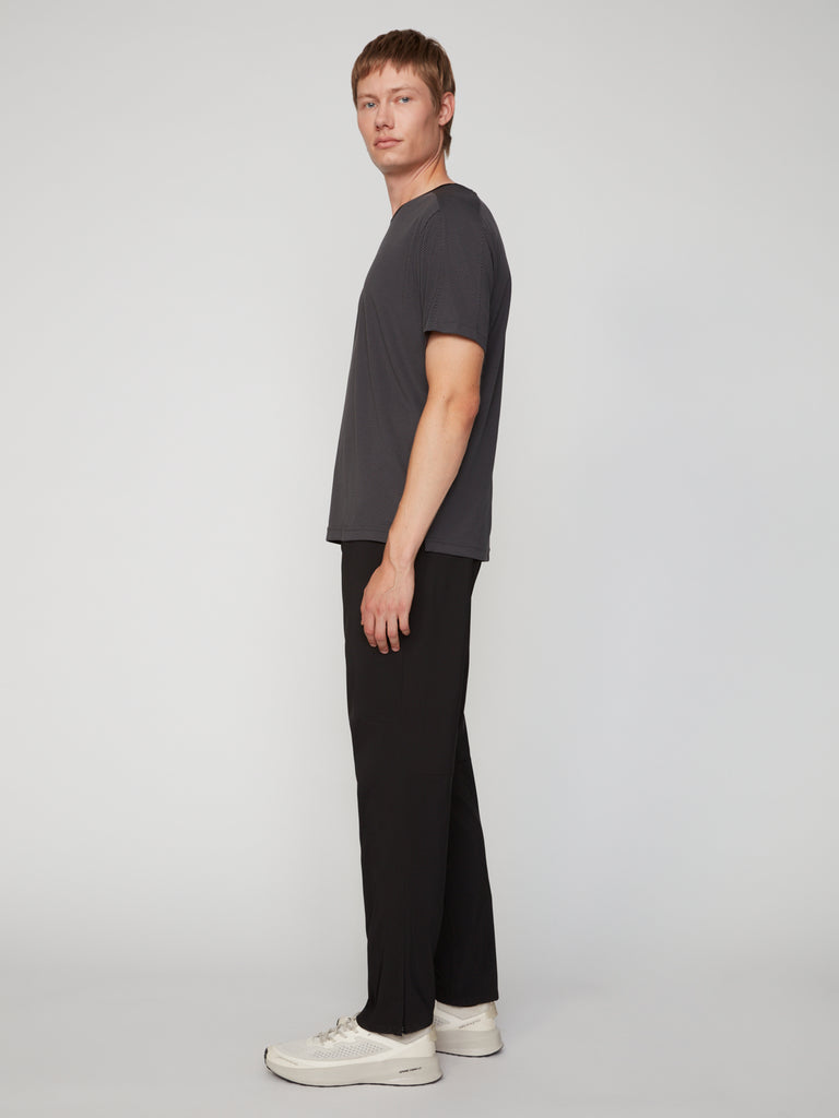 A person with short light brown hair, in a dark gray t-shirt, TECH APPAREL MEN’S PULL-ON TRAINING PANT (RELAXED TAPERED FIT), and white sneakers stands in profile against a plain light gray background.