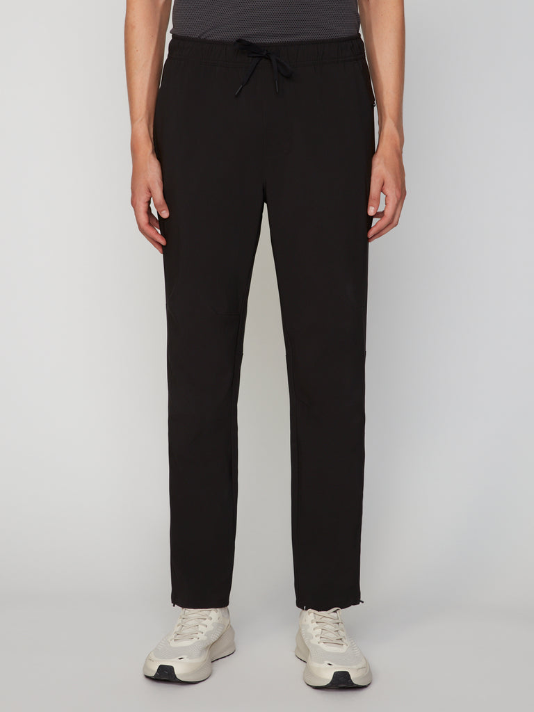 A person wears TECH APPAREL MEN’S PULL-ON TRAINING PANT, RELAXED TAPERED FIT in black with white sneakers, standing against a plain light background. Only their lower torso and legs are visible; hands rest by their sides.