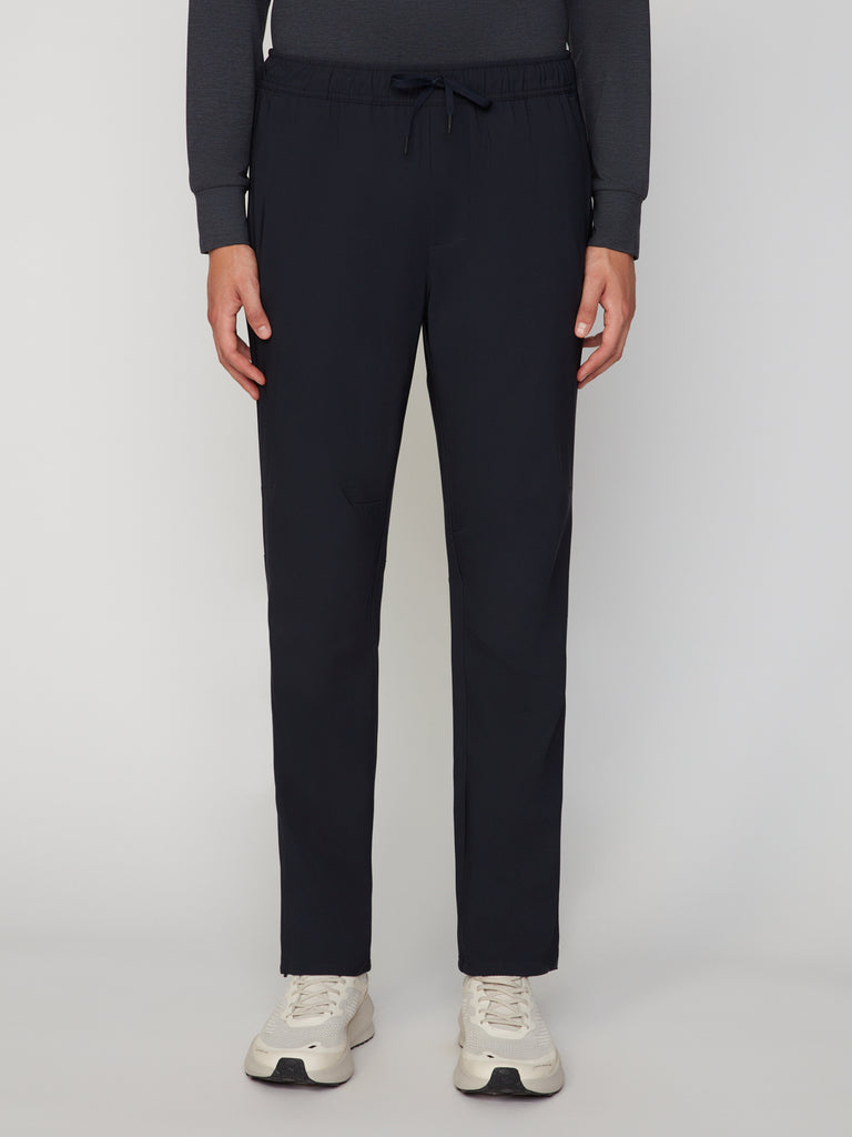 A person, shown from the shoulders down, wears TECH APPAREL MEN’S PULL-ON TRAINING PANT, RELAXED TAPERED FIT in black with a dark long-sleeve shirt and white sneakers, standing against a plain white background.