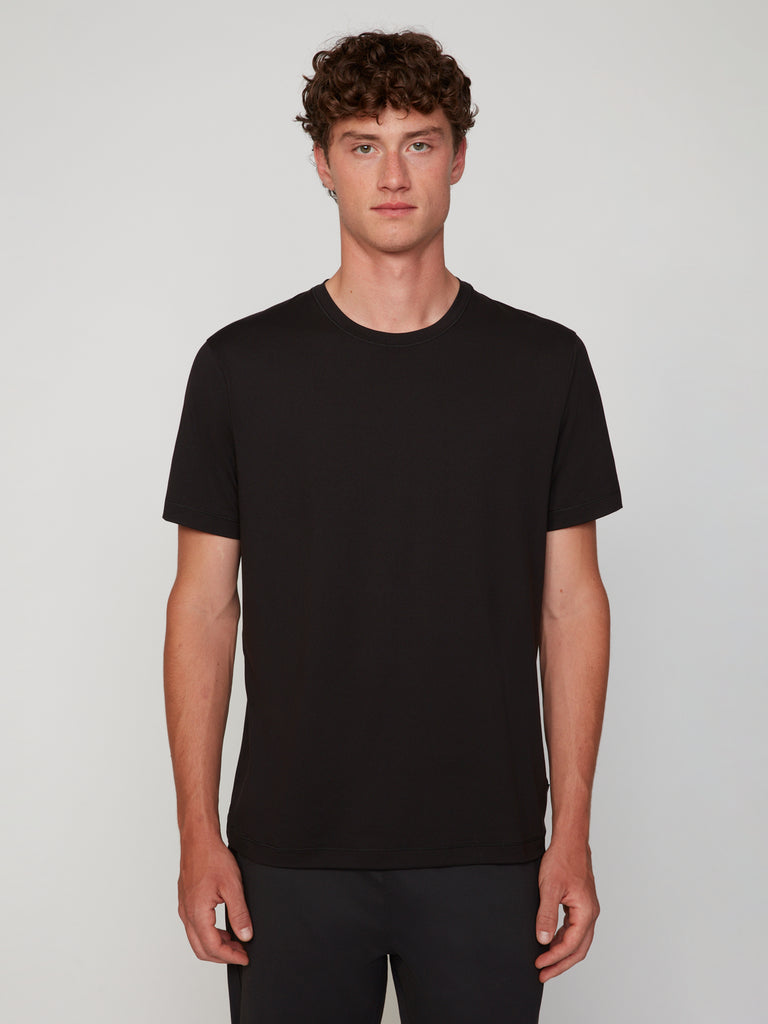 A young man with curly brown hair stands against a light background, facing forward in a neutral expression. He wears TECH APPAREL’s MEN’S PEACH:TECH CLASSIC FIT CREW NECK T-SHIRT and black pants.