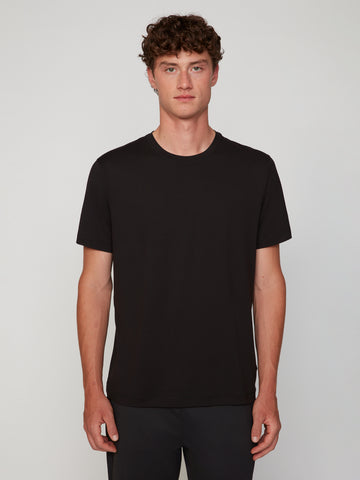 A young man with curly brown hair stands against a light background, facing forward in a neutral expression. He wears TECH APPAREL’s MEN’S PEACH:TECH CLASSIC FIT CREW NECK T-SHIRT and black pants.