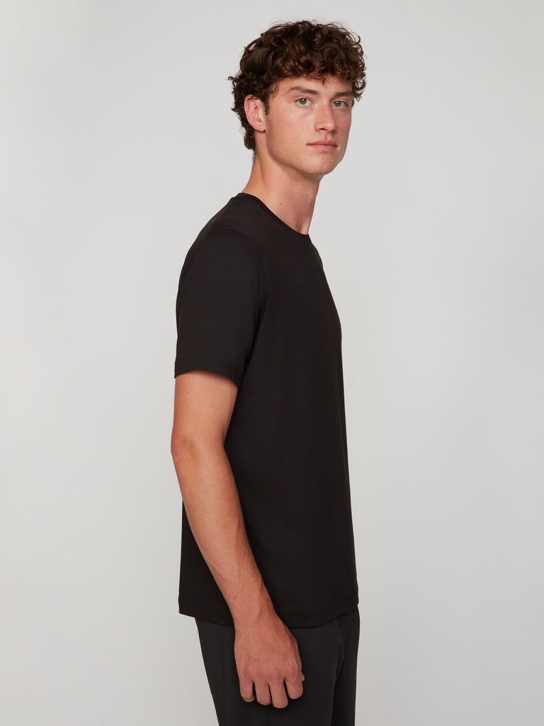 A young man with curly brown hair stands in profile against a plain light gray background, wearing the TECH APPAREL MEN’S PEACH:TECH CLASSIC FIT CREW NECK T-SHIRT in black and dark pants.