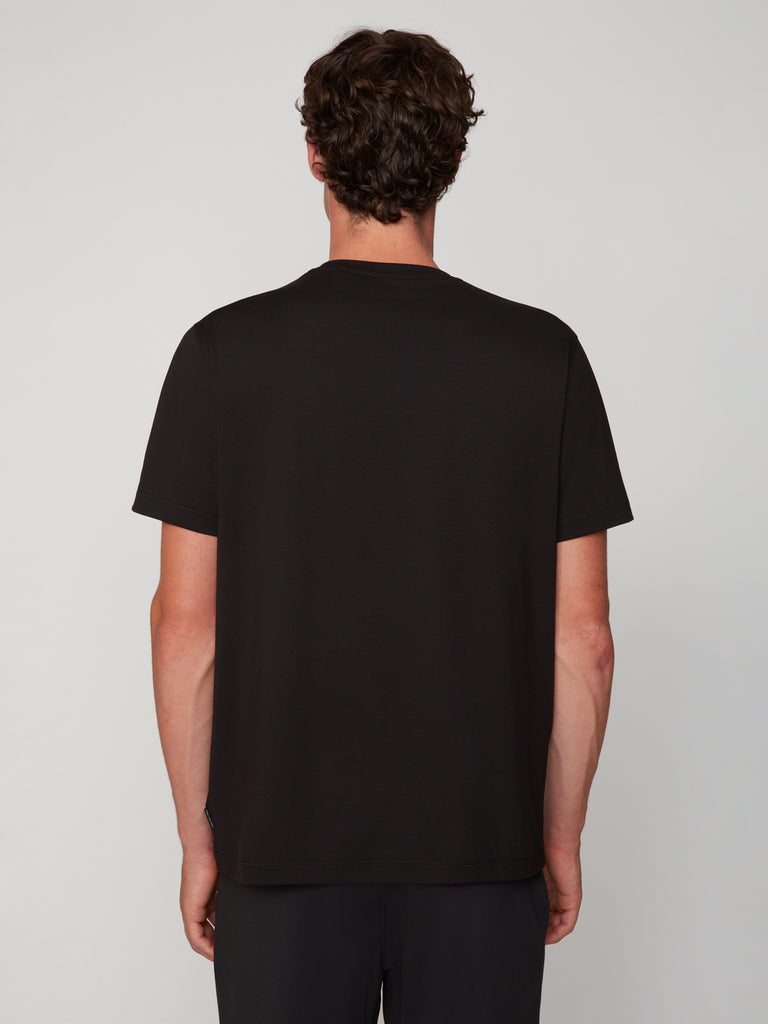 A man with short, curly brown hair stands with his back to the camera, wearing a TECH APPAREL MEN’S PEACH:TECH CLASSIC FIT CREW NECK T-SHIRT and black pants against a light gray background.