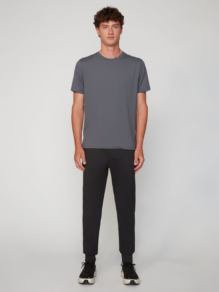 A young man with curly brown hair wears the TECH APPAREL MEN’S PEACH:TECH CLASSIC FIT CREW NECK T-SHIRT in gray, paired with black joggers and sneakers, standing against a plain light gray background.