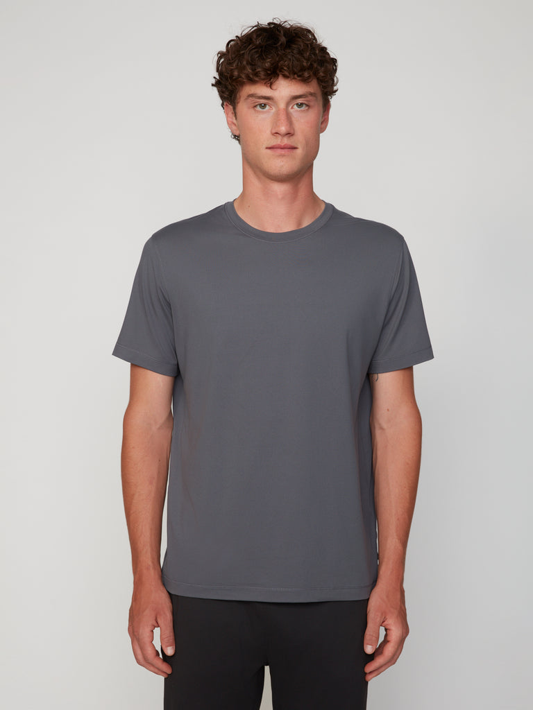A young man with curly brown hair faces forward against a light gray background, wearing the TECH APPAREL MEN’S PEACH:TECH CLASSIC FIT CREW NECK T-SHIRT in gray and black pants.