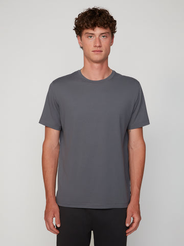 A young man with curly brown hair faces forward against a light gray background, wearing the TECH APPAREL MEN’S PEACH:TECH CLASSIC FIT CREW NECK T-SHIRT in gray and black pants.