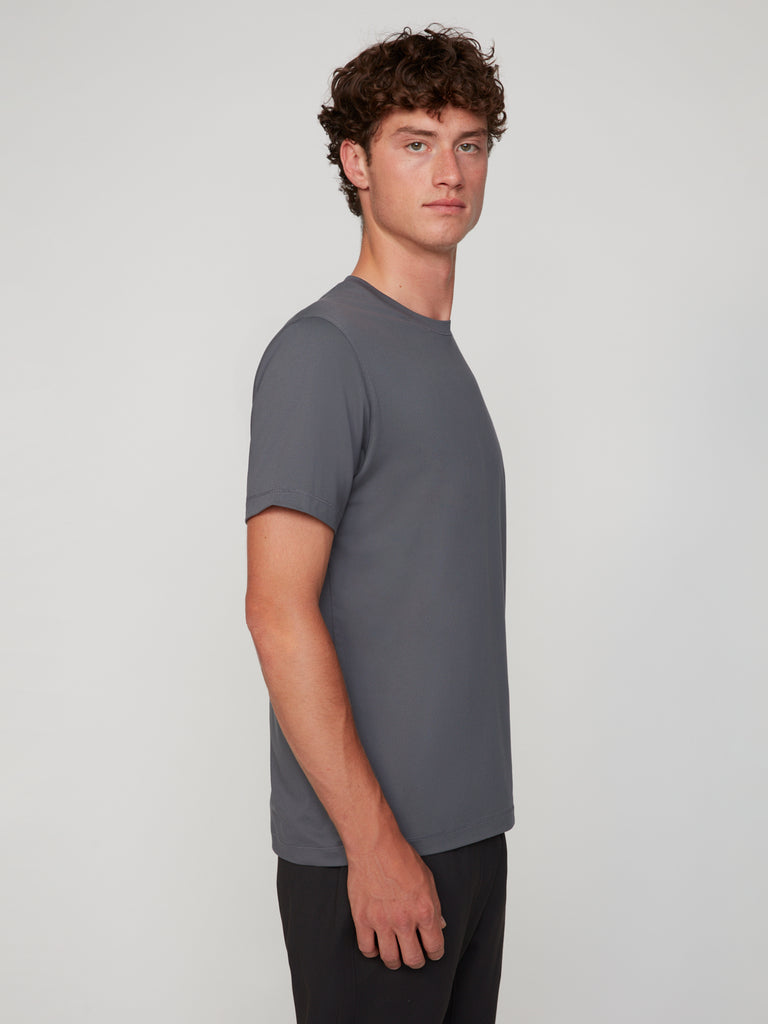 A young man with curly brown hair stands in profile against a plain background, wearing TECH APPARELs MEN’S PEACH:TECH CLASSIC FIT CREW NECK T-SHIRT in gray and dark pants, looking straight ahead with a neutral expression.