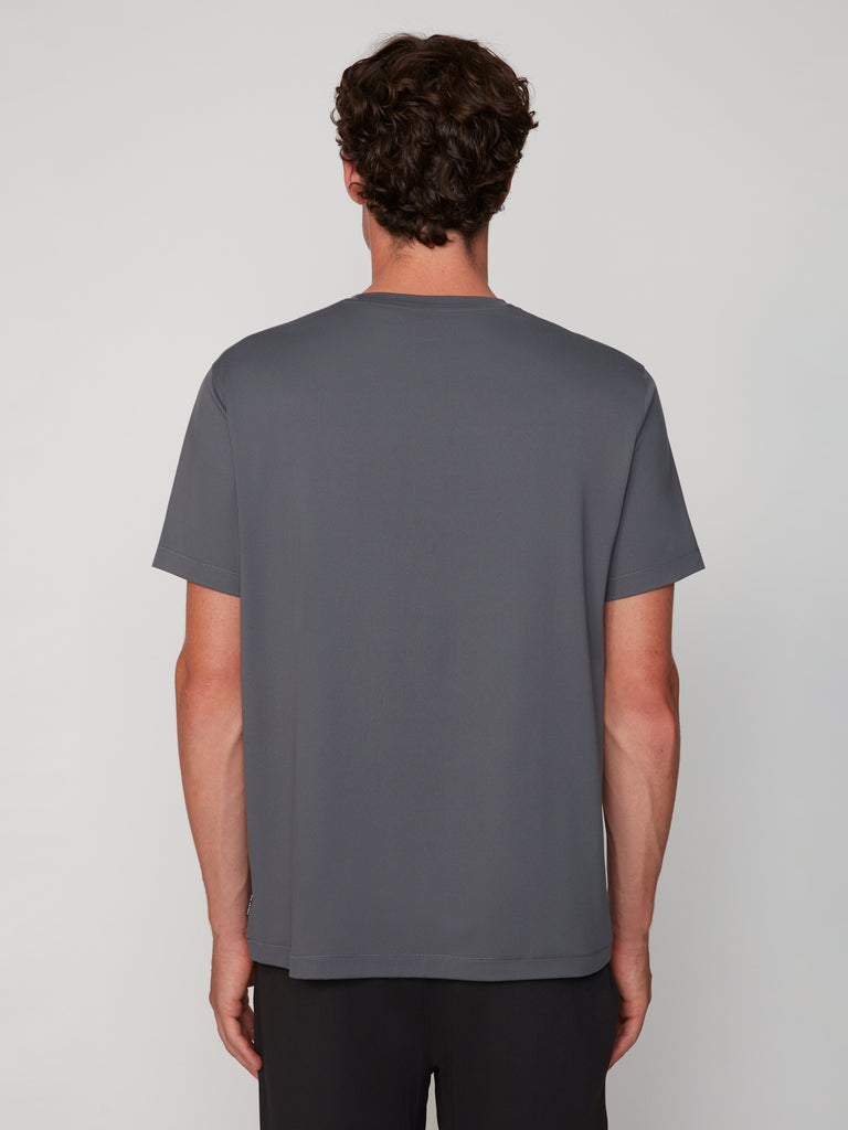 A man with short curly hair stands with his back to the camera, wearing the TECH APPAREL MEN’S PEACH:TECH CLASSIC FIT CREW NECK T-SHIRT in plain gray and dark pants against a light gray background.