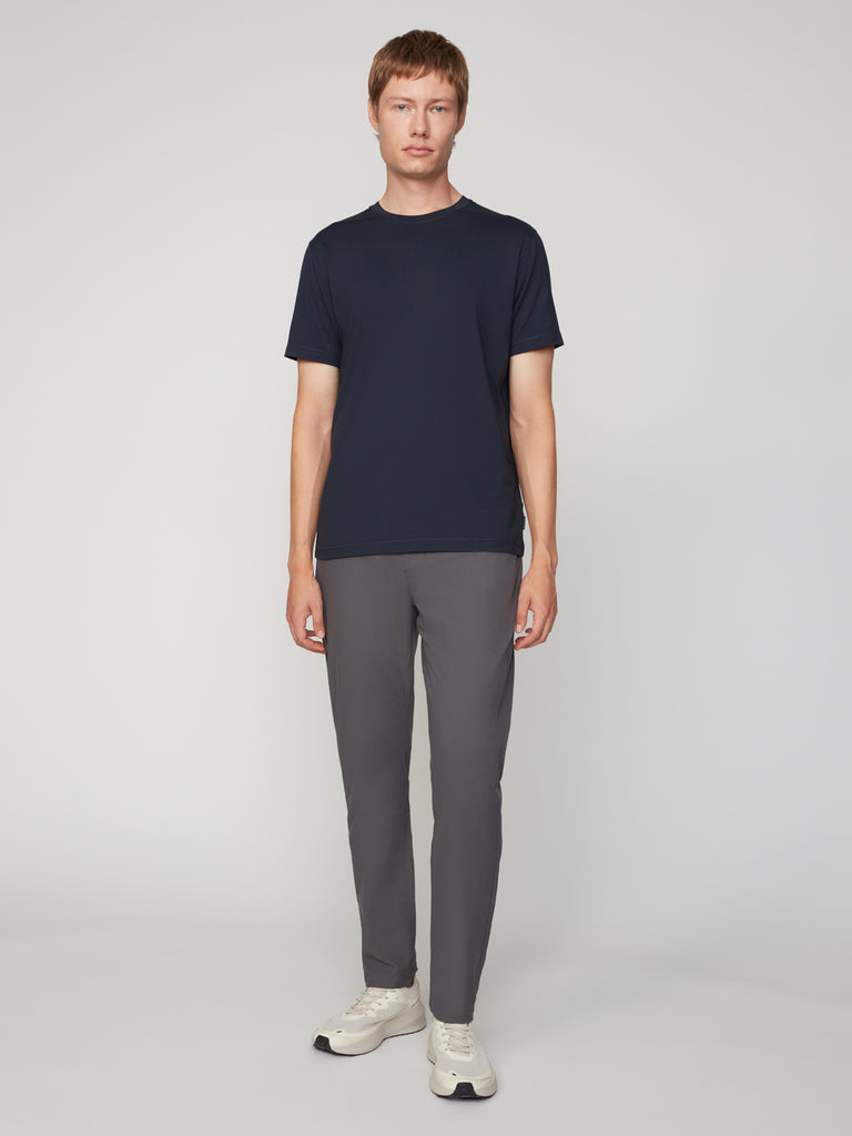 A man with short, light brown hair stands facing forward against a light gray background, wearing a TECH APPAREL MEN’S PEACH:TECH CLASSIC FIT CREW NECK T-SHIRT in navy blue, gray pants, and white sneakers.