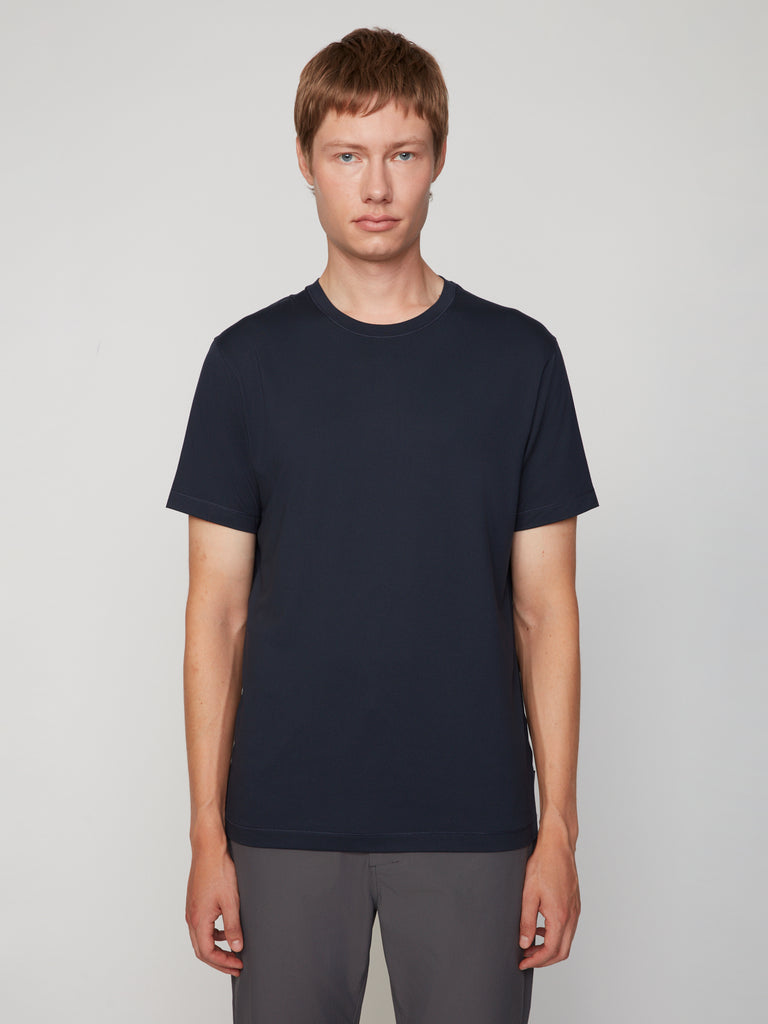 A person with short light brown hair stands against a plain background, wearing TECH APPAREL’s MEN’S PEACH:TECH CLASSIC FIT CREW NECK T-SHIRT in dark navy and gray pants, looking at the camera with a neutral expression.