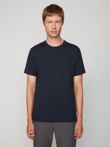 A person with short light brown hair stands against a plain background, wearing TECH APPAREL’s MEN’S PEACH:TECH CLASSIC FIT CREW NECK T-SHIRT in dark navy and gray pants, looking at the camera with a neutral expression.