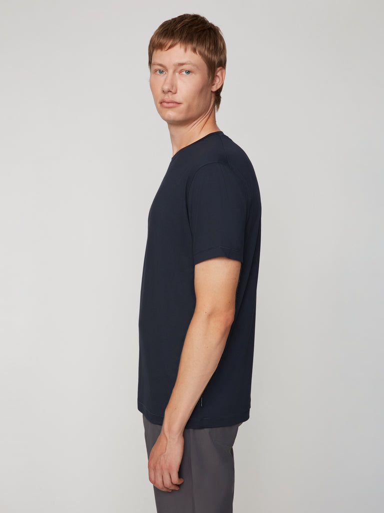 A person with short light brown hair stands sideways, wearing the TECH APPAREL MEN’S PEACH:TECH CLASSIC FIT CREW NECK T-SHIRT in dark navy and gray pants, looking calmly at the camera against a plain light background.