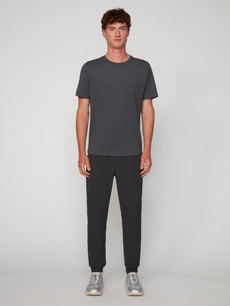 A man with short curly hair stands facing forward against a plain background, wearing TECH APPAREL’S MEN’S CREW NECK TEE WITH MIXED JACQUARD STRIPE, paired with dark pants and grey sneakers for lightweight comfort.