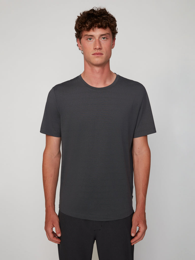 A young man with short curly brown hair wears the TECH APPAREL MEN’S CREW NECK TEE WITH MIXED JACQUARD STRIPE in dark gray and dark pants, standing against a light gray background and looking straight at the camera.
