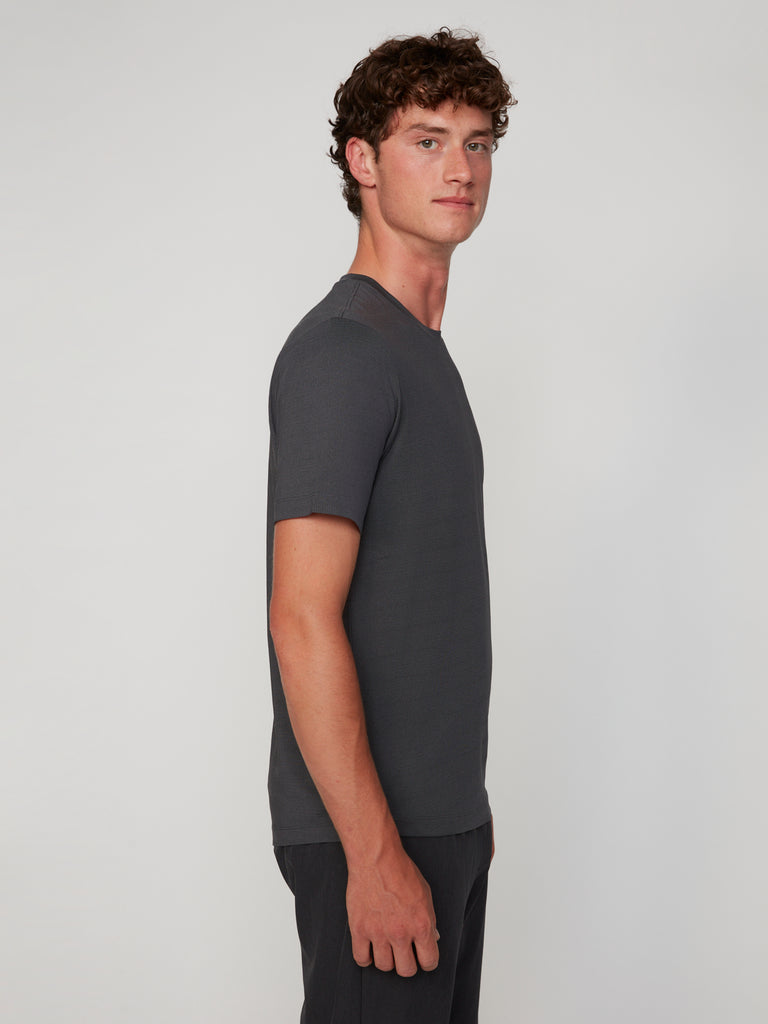 A young man with curly brown hair stands in profile, facing left, wearing a TECH APPAREL MEN’S CREW NECK TEE WITH MIXED JACQUARD STRIPE and matching pants, offering lightweight comfort against a plain light grey background.