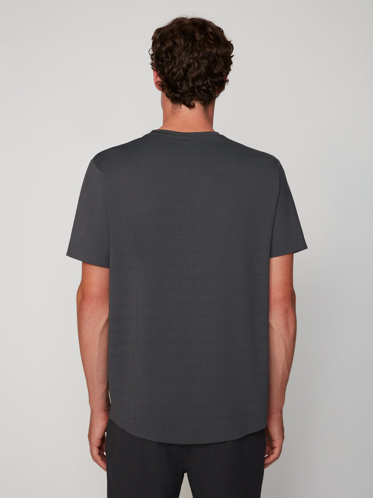 A person with short, curly brown hair faces away from the camera, wearing TECH APPAREL’s MEN’S CREW NECK TEE WITH MIXED JACQUARD STRIPE and dark pants against a plain light grey background.