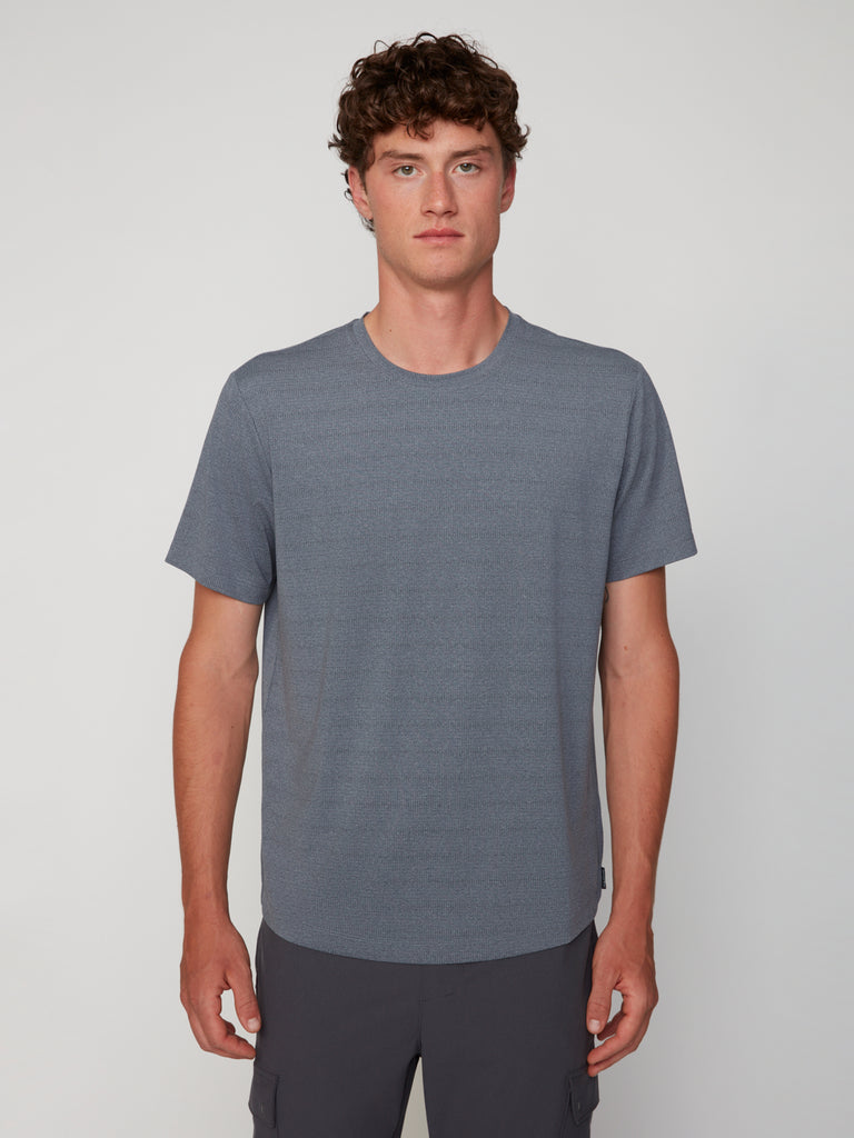 A young man with curly brown hair wears a TECH APPAREL MEN’S CREW NECK TEE WITH MIXED JACQUARD STRIPE in soft gray, standing facing forward against a plain, light background.