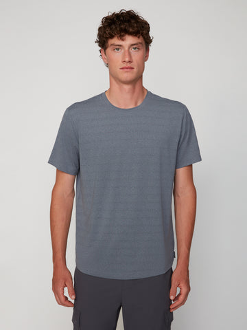 A young man with curly brown hair wears a TECH APPAREL MEN’S CREW NECK TEE WITH MIXED JACQUARD STRIPE in soft gray, standing facing forward against a plain, light background.