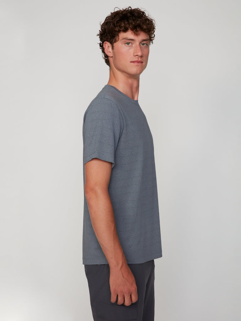 A young man with curly brown hair stands in profile against a light background, wearing the TECH APPAREL MEN’S CREW NECK TEE WITH MIXED JACQUARD STRIPE and dark shorts, looking forward with a neutral expression.