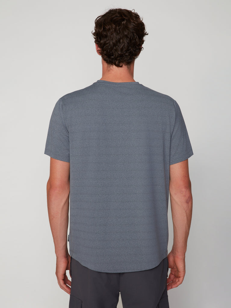 A man with short brown hair faces away from the camera, wearing the TECH APPAREL MEN’S CREW NECK TEE WITH MIXED JACQUARD STRIPE and dark gray pants against a plain light background.