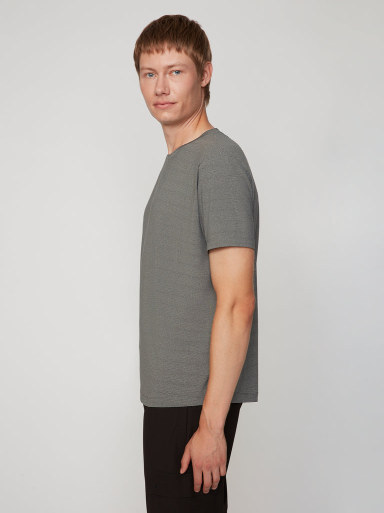 A person with short light brown hair stands in profile, facing left, wearing the TECH APPAREL MEN’S CREW NECK TEE WITH MIXED JACQUARD STRIPE for lightweight comfort and black pants against a plain light background.