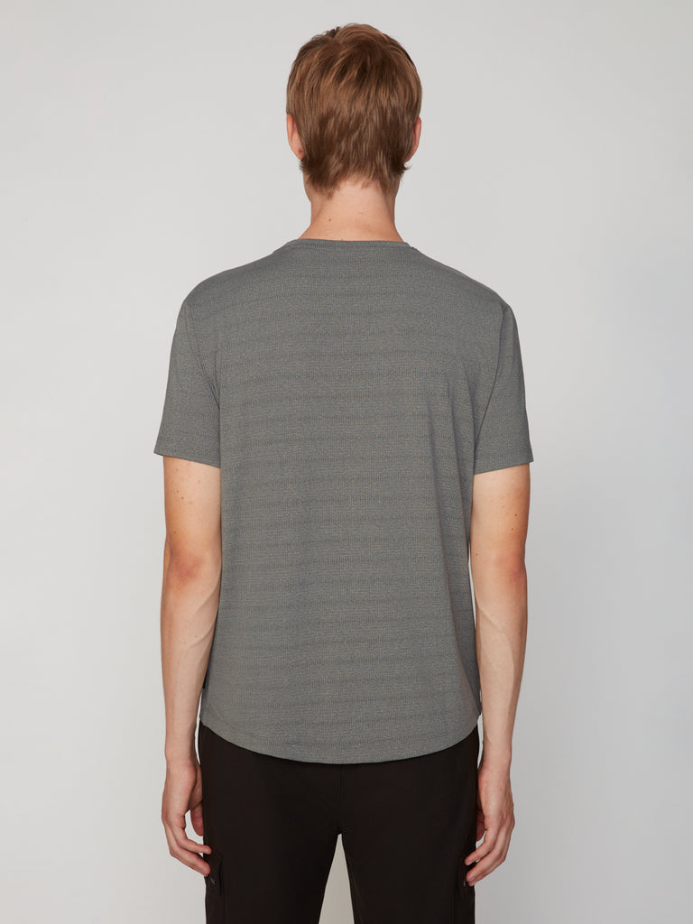 A person with short light brown hair is seen from behind, wearing a gray TECH APPAREL MEN’S CREW NECK TEE WITH MIXED JACQUARD STRIPE and black pants, standing against a plain, light-colored background.