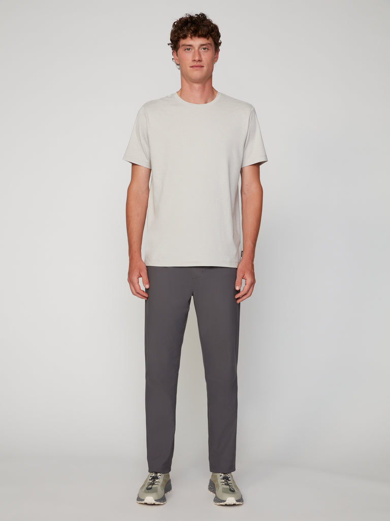 A man with short curly hair faces forward, wearing TECH APPAREL’s MEN’S CREW NECK T-SHIRT IN BRUSHED SPACE-DYED JERSEY, paired with dark gray pants and gray sneakers, against a plain light background.