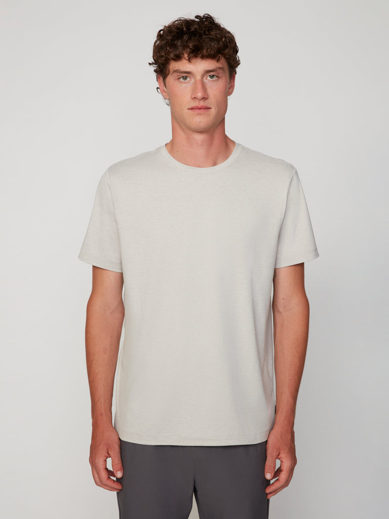 A person with short curly brown hair wears the TECH APPAREL MEN’S CREW NECK T-SHIRT IN BRUSHED SPACE-DYED JERSEY in light beige and dark pants, standing facing forward against a plain light background.