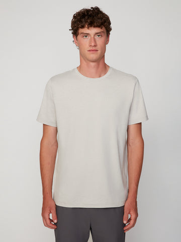 A person with short curly brown hair wears the TECH APPAREL MEN’S CREW NECK T-SHIRT IN BRUSHED SPACE-DYED JERSEY in light beige and dark pants, standing facing forward against a plain light background.