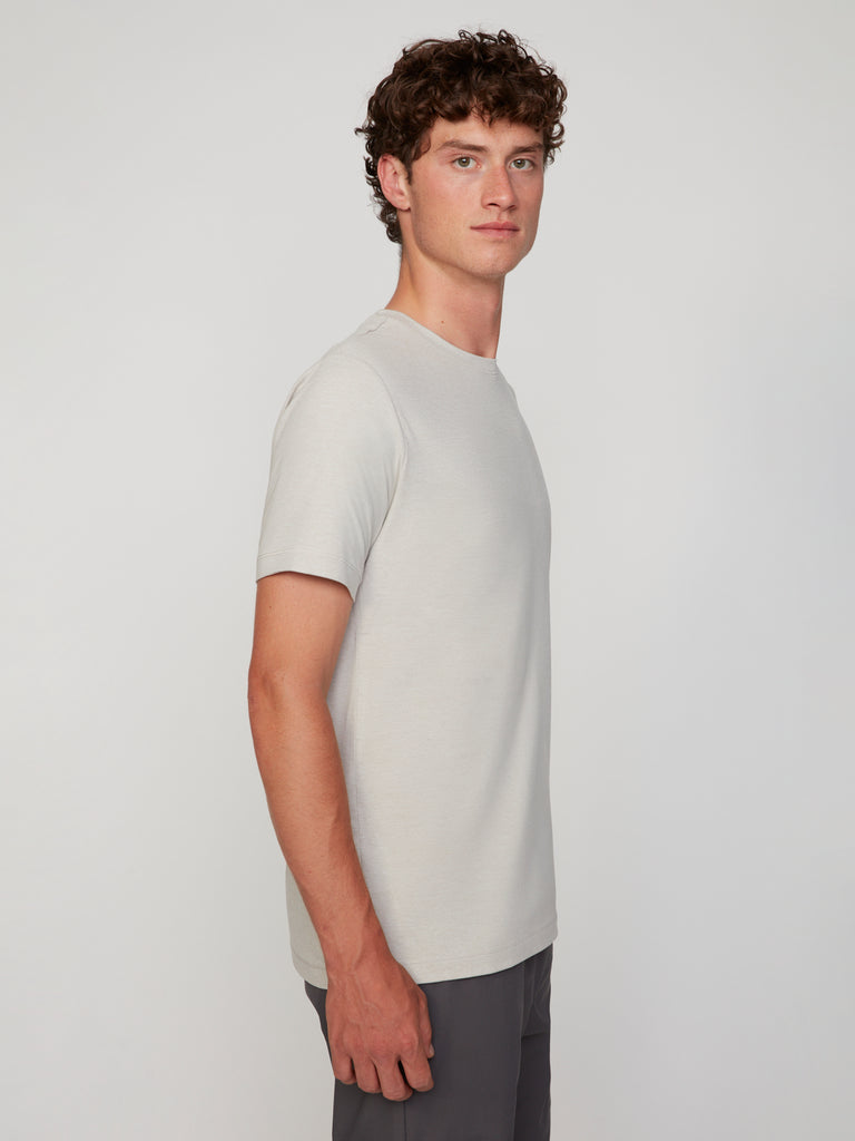 A young man with curly brown hair stands sideways, facing left, wearing the TECH APPAREL MEN’S CREW NECK T-SHIRT IN BRUSHED SPACE-DYED JERSEY and dark pants against a plain light background.