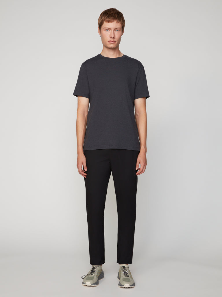 A person stands facing forward against a light gray background, wearing TECH APPAREL’s MEN’S CREW NECK T-SHIRT IN BRUSHED SPACE-DYED JERSEY, black pants, and light gray sneakers, with arms at their sides.