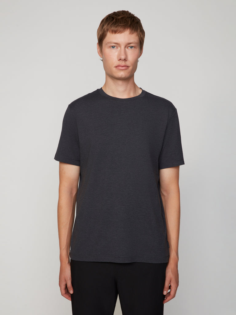 A person with short light brown hair stands facing forward, wearing a TECH APPAREL MEN’S CREW NECK T-SHIRT IN BRUSHED SPACE-DYED JERSEY in dark gray and black pants, set against a neutral light gray background.