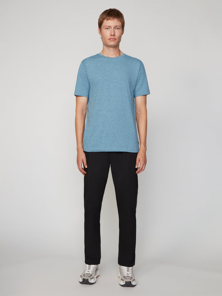 A person stands facing forward against a plain light background, wearing a TECH APPAREL MEN’S CREW NECK T-SHIRT IN BRUSHED SPACE-DYED JERSEY, black pants, and gray sneakers.