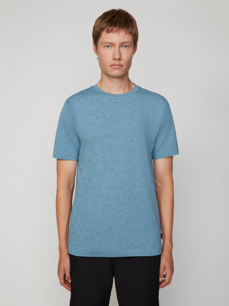 A person with short light brown hair wears the TECH APPAREL MEN’S CREW NECK T-SHIRT IN BRUSHED SPACE-DYED JERSEY in light blue and black pants, standing facing forward against a plain light gray background.
