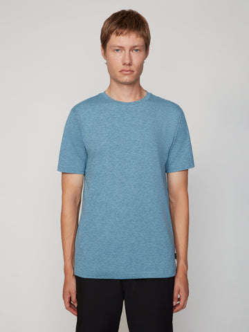 A person with short light brown hair wears the TECH APPAREL MEN’S CREW NECK T-SHIRT IN BRUSHED SPACE-DYED JERSEY in light blue and black pants, standing facing forward against a plain light gray background.