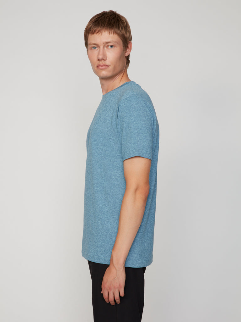A man with short light brown hair models the TECH APPAREL MEN’S CREW NECK T-SHIRT IN BRUSHED SPACE-DYED JERSEY in blue with black pants, standing sideways and looking at the camera against a plain light background.