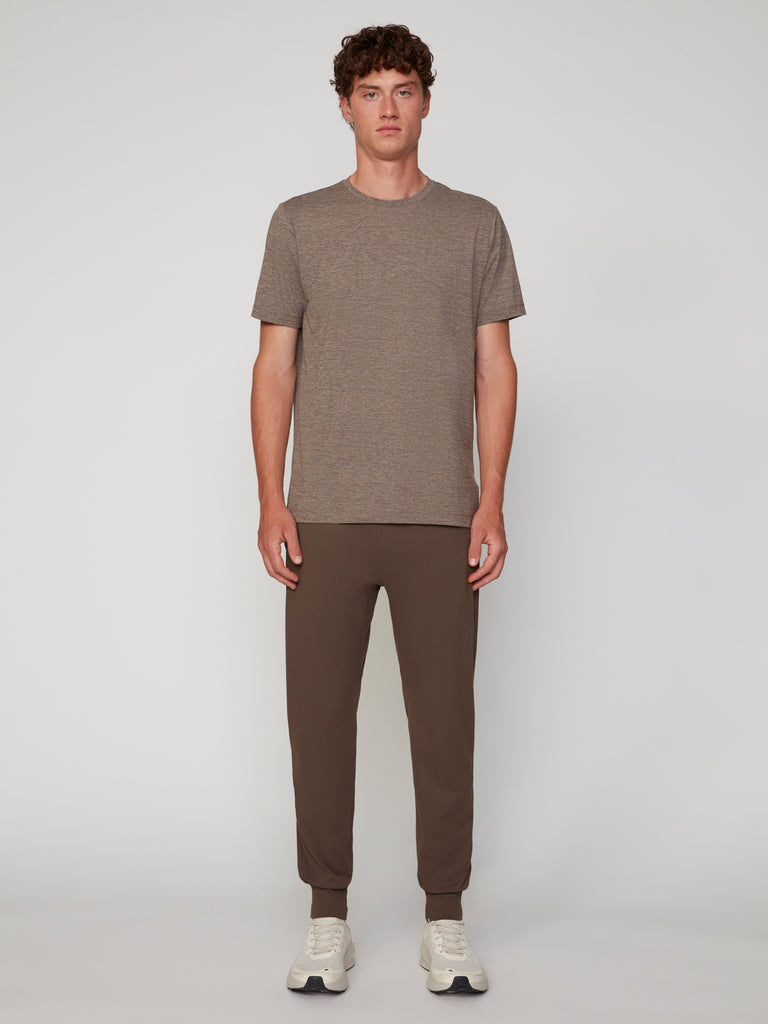 A man faces forward against a plain background, wearing TECH APPAREL’s MEN’S CREW NECK T-SHIRT IN BRUSHED SPACE-DYED JERSEY in taupe, paired with brown jogger pants and white sneakers.