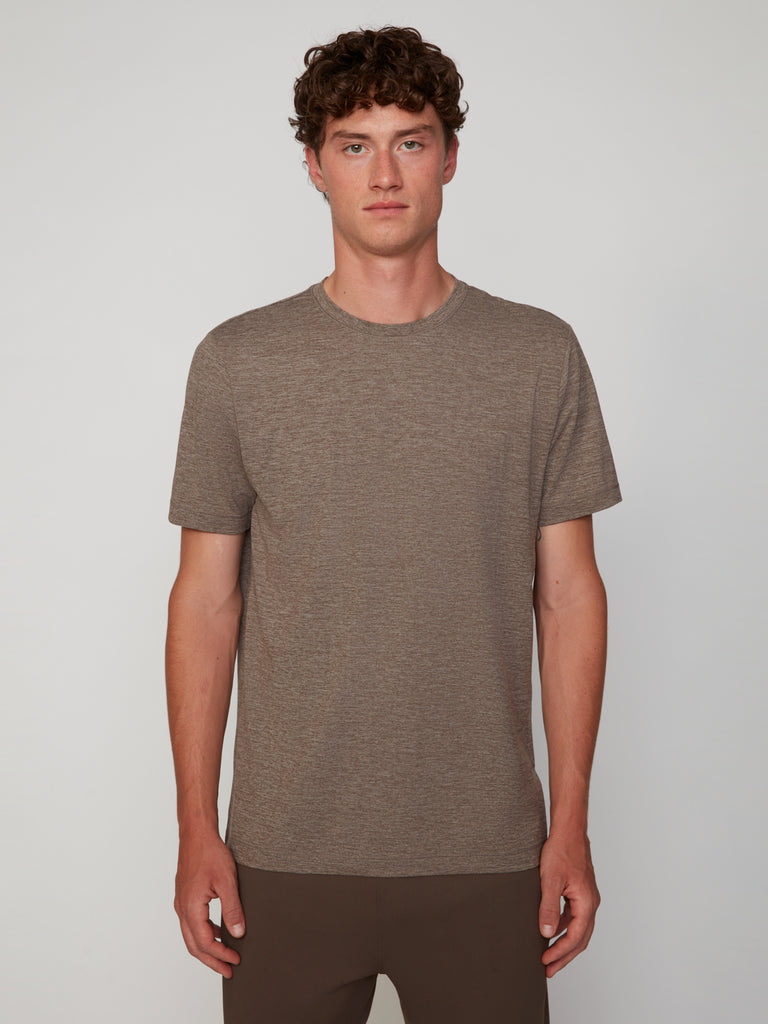A young man with curly brown hair wears a TECH APPAREL MEN’S CREW NECK T-SHIRT IN BRUSHED SPACE-DYED JERSEY, standing facing forward against a simple light gray background.