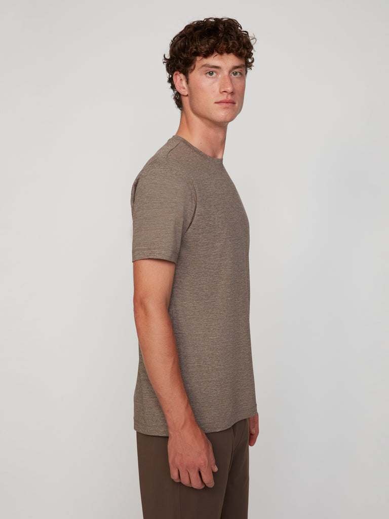 A young man with curly brown hair stands in profile against a light gray background, wearing TECH APPAREL’s MEN’S CREW NECK T-SHIRT IN BRUSHED SPACE-DYED JERSEY, paired with brown pants.