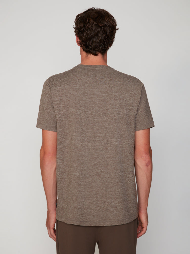 A person with short, curly brown hair stands with their back to the camera, wearing TECH APPAREL’s MEN’S CREW NECK T-SHIRT IN BRUSHED SPACE-DYED JERSEY and brown pants against a plain light background.
