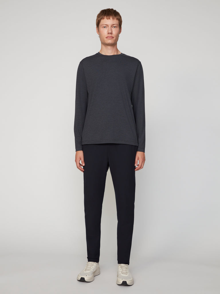 A person stands facing forward against a plain background, wearing the TECH APPAREL MEN’S LONG SLEEVE CREW NECK IN BRUSHED SPACE-DYED JERSEY, paired with dark pants and white sneakers.