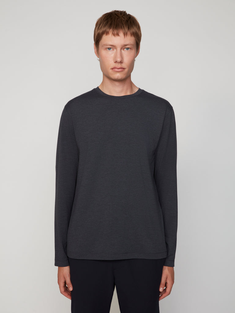 A person with short light brown hair wears TECH APPAREL’s MEN’S LONG SLEEVE CREW NECK IN BRUSHED SPACE-DYED JERSEY and black pants, standing against a plain light background.