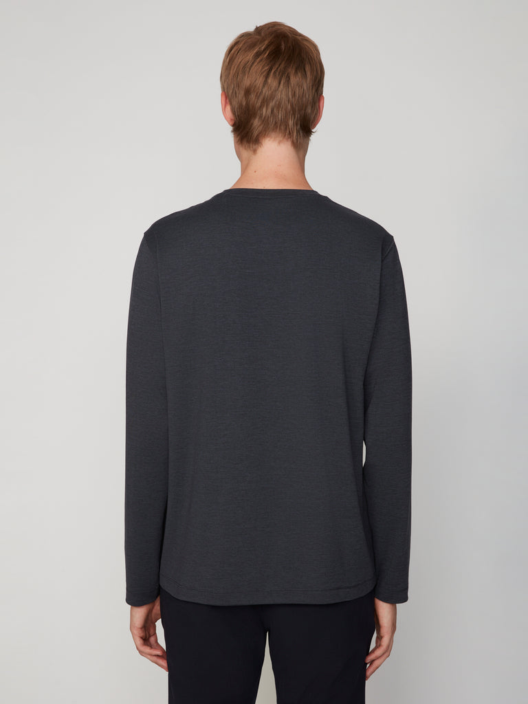 A person with short light brown hair is shown from the back wearing TECH APPAREL’s MEN’S LONG SLEEVE CREW NECK IN BRUSHED SPACE-DYED JERSEY and black pants, standing against a plain light gray background.