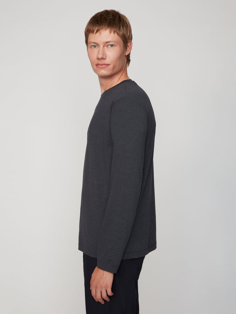 A man with short light brown hair, wearing TECH APPAREL’s MEN’S LONG SLEEVE CREW NECK IN BRUSHED SPACE-DYED JERSEY and dark pants, stands sideways and looks at the camera against a plain light background.