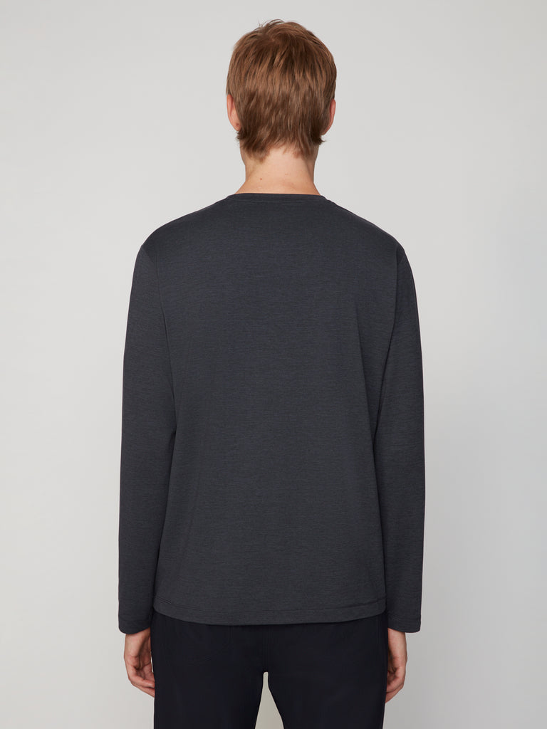 A person with short light brown hair is seen from behind, wearing TECH APPAREL’s MEN’S LONG SLEEVE CREW NECK IN BRUSHED SPACE-DYED JERSEY and black pants, standing against a plain light gray background.