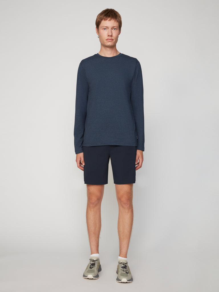 A person stands facing forward against a plain background, wearing TECH APPAREL’s MEN’S LONG SLEEVE CREW NECK IN BRUSHED SPACE-DYED JERSEY in navy, navy shorts, white socks, and gray sneakers.