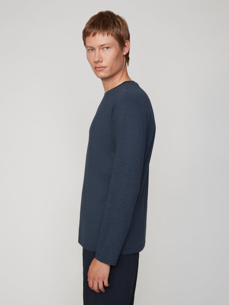 A man with short light brown hair models the TECH APPAREL MEN’S LONG SLEEVE CREW NECK IN BRUSHED SPACE-DYED JERSEY in dark blue, paired with dark pants, standing sideways and looking at the camera against a plain light background.