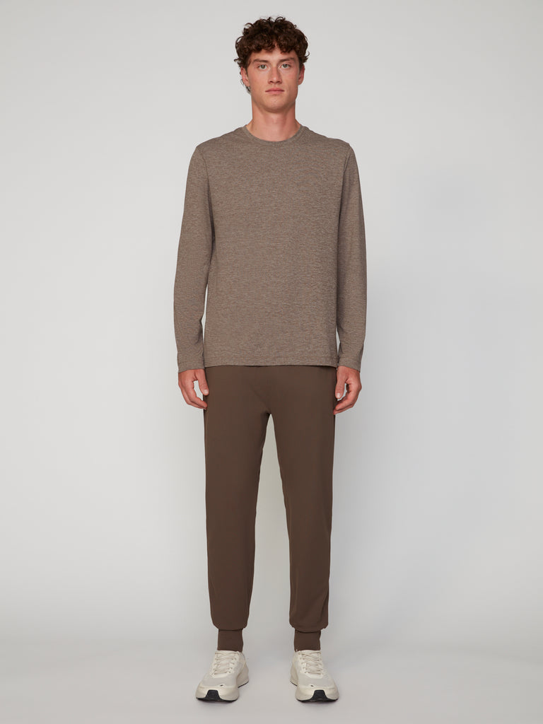 A young man stands against a light gray background wearing TECH APPARELs MEN’S LONG SLEEVE CREW NECK IN BRUSHED SPACE-DYED JERSEY in taupe, paired with brown jogger pants and white sneakers. He faces forward with a neutral expression.