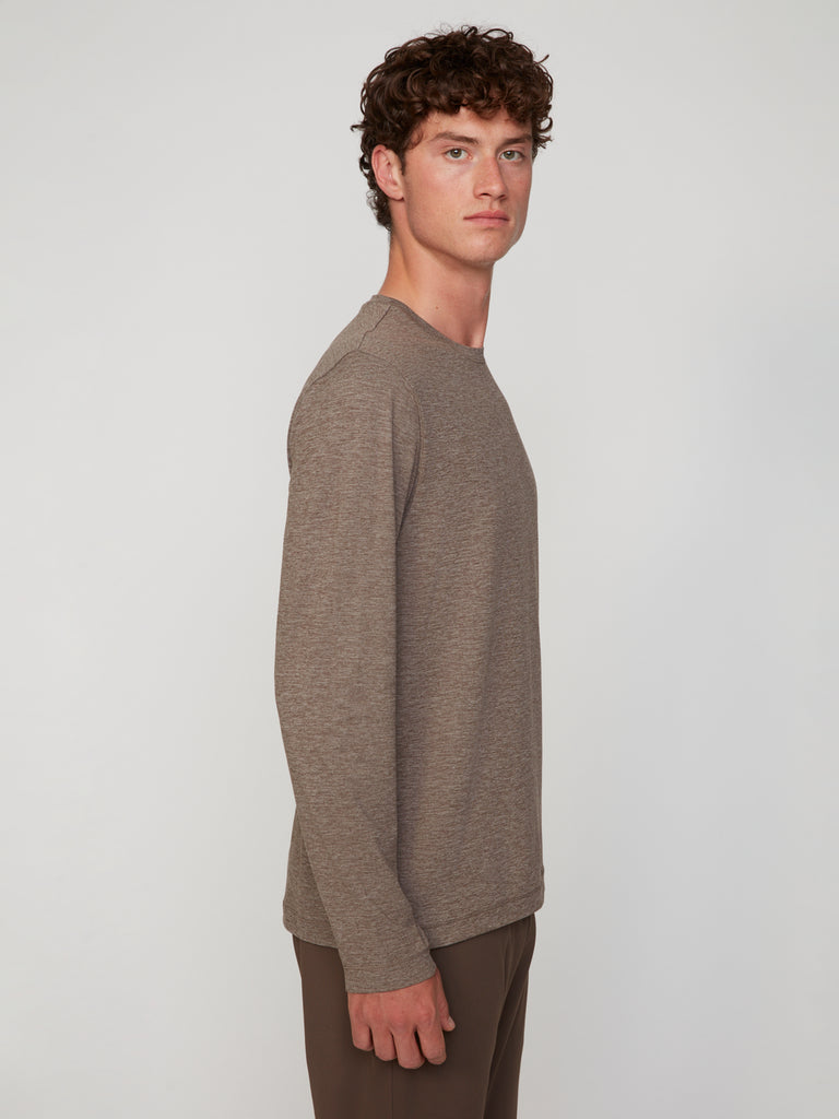 A young man with curly brown hair models the TECH APPAREL MEN’S LONG SLEEVE CREW NECK IN BRUSHED SPACE-DYED JERSEY with light brown pants, highlighting its breathable performance against a plain, light gray background.