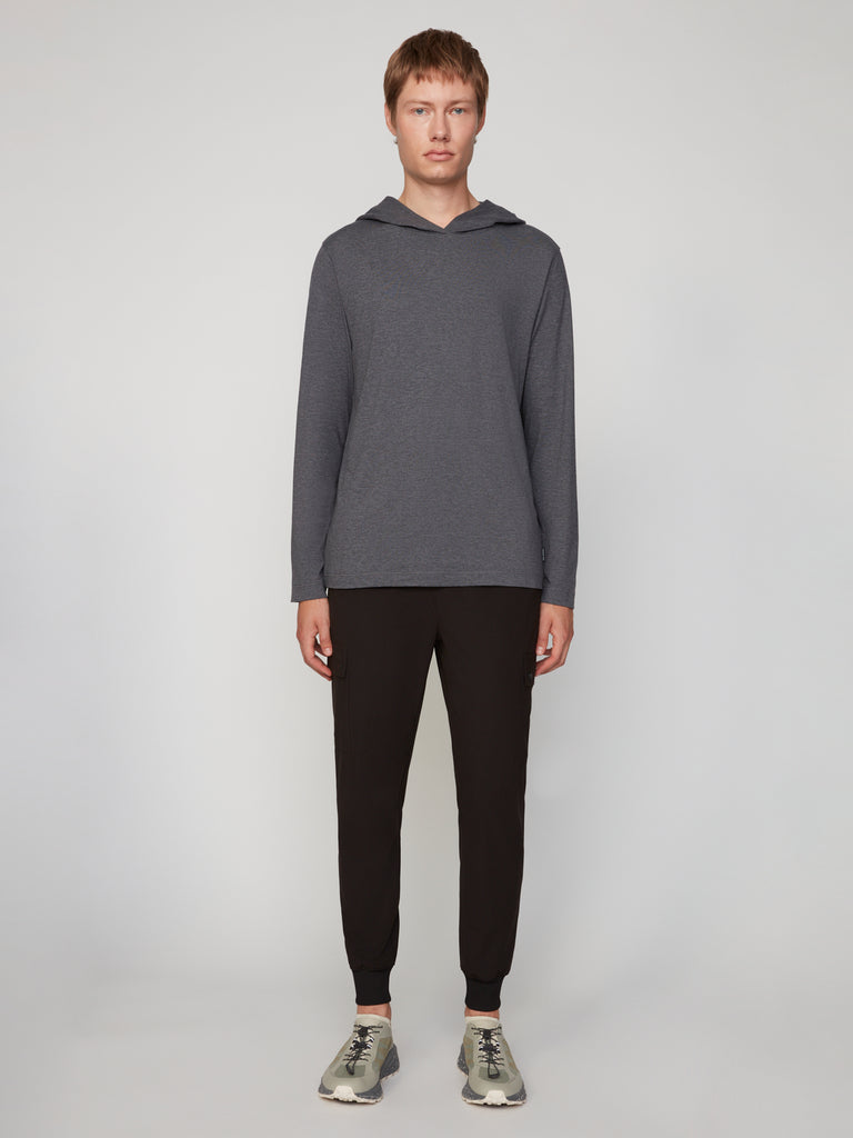 A person faces forward against a light gray background, wearing TECH APPAREL’s MEN’S LONG SLEEVE HOODIE IN BRUSHED SPACE-DYED JERSEY with black jogger pants and gray sneakers—ideal for comfortable, versatile casual style.